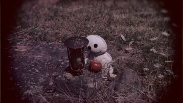 Das Wesen sieht an einem kleinen Grab, einer Grableuchte die auf einem Stein steht und neben ihr ein roter Apfel liegt. Das Wesen lächelt