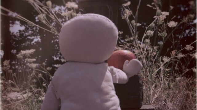 Das Wesen von hinten photograhiert als es einen roten Apfel auf den Grabstein legt auf dem eine Grableuchte steht. Um das Grab wachsen verwilderte Blumen.