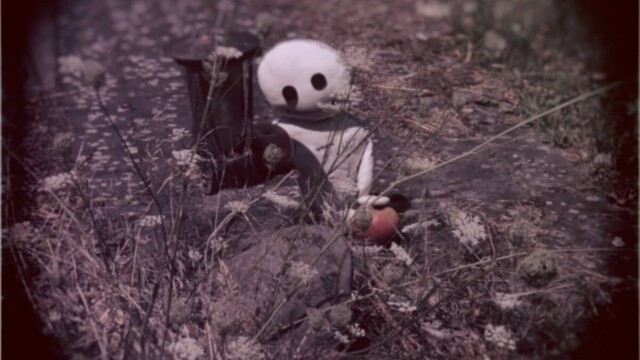 Das Wesen sieht an einem kleinen Grab, einer Grableuchte die auf einem Stein steht und neben ihr ein roter Apfel liegt. Das Wesen lächelt