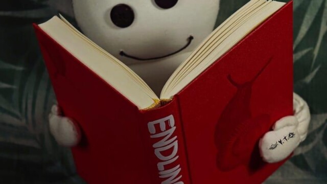 Das Wesen sitzt vor dem aufgeschlagenem Buch von Jasmin Schreiber: Endling. Es scheint lächelnd darin zu lesen.