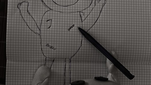 Das Wesen liegt auf dem Tisch auf einem ausgebreiteten Schreibblock. Auf dem Karopapier wurde ein Wesenabbild mit Kugelschreiber gemalt. Den Kugelschreiber hält das Wesen in einer Pfote. Das Bild wurde von oben fotografiert.