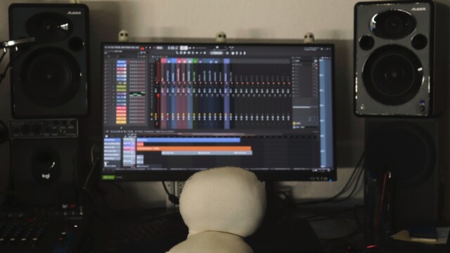 Das Wesen von hinten fotografiert sitzt an einem großen Tisch mit Computermonitor, Lautsprecherboxen, Mischpult und weiteren Musikequipment. Auf dem Monitor ist ein Musiksequencerprorgramm geöffnet. Auf dem Monitorrand stehen drei Geisterfiguren. Auf der Lautsprecherbox linke Seite sitzt eine Kodamafigur.